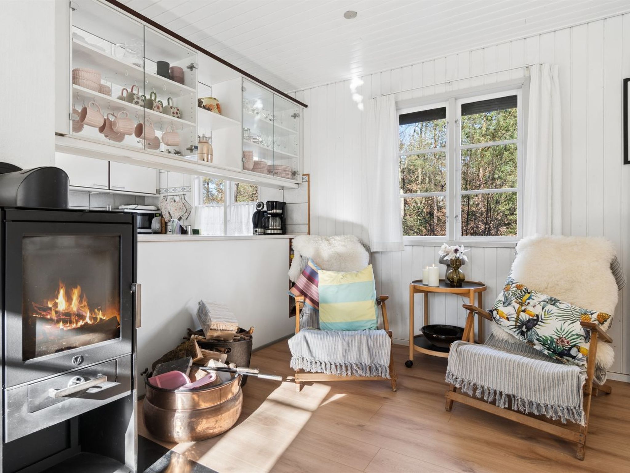 Ett mysigt rum med en vedeldad kamin, en fåtölj med en färgglad kudde och en hylla med diverse köksartiklar.