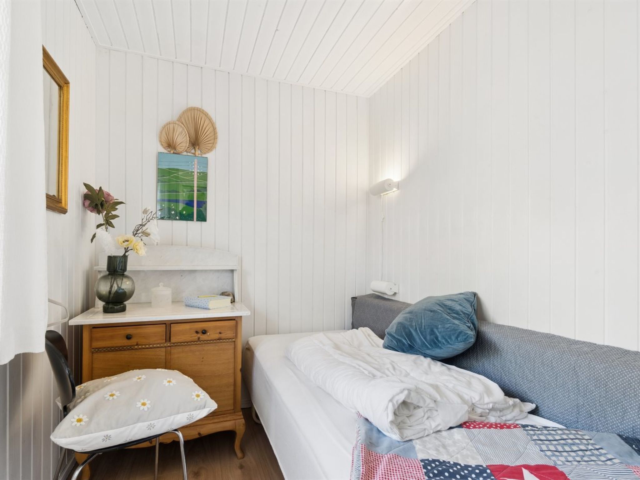 A bedroom with a bed, a desk, a chair, a wall-mounted lamp, and a framed picture.