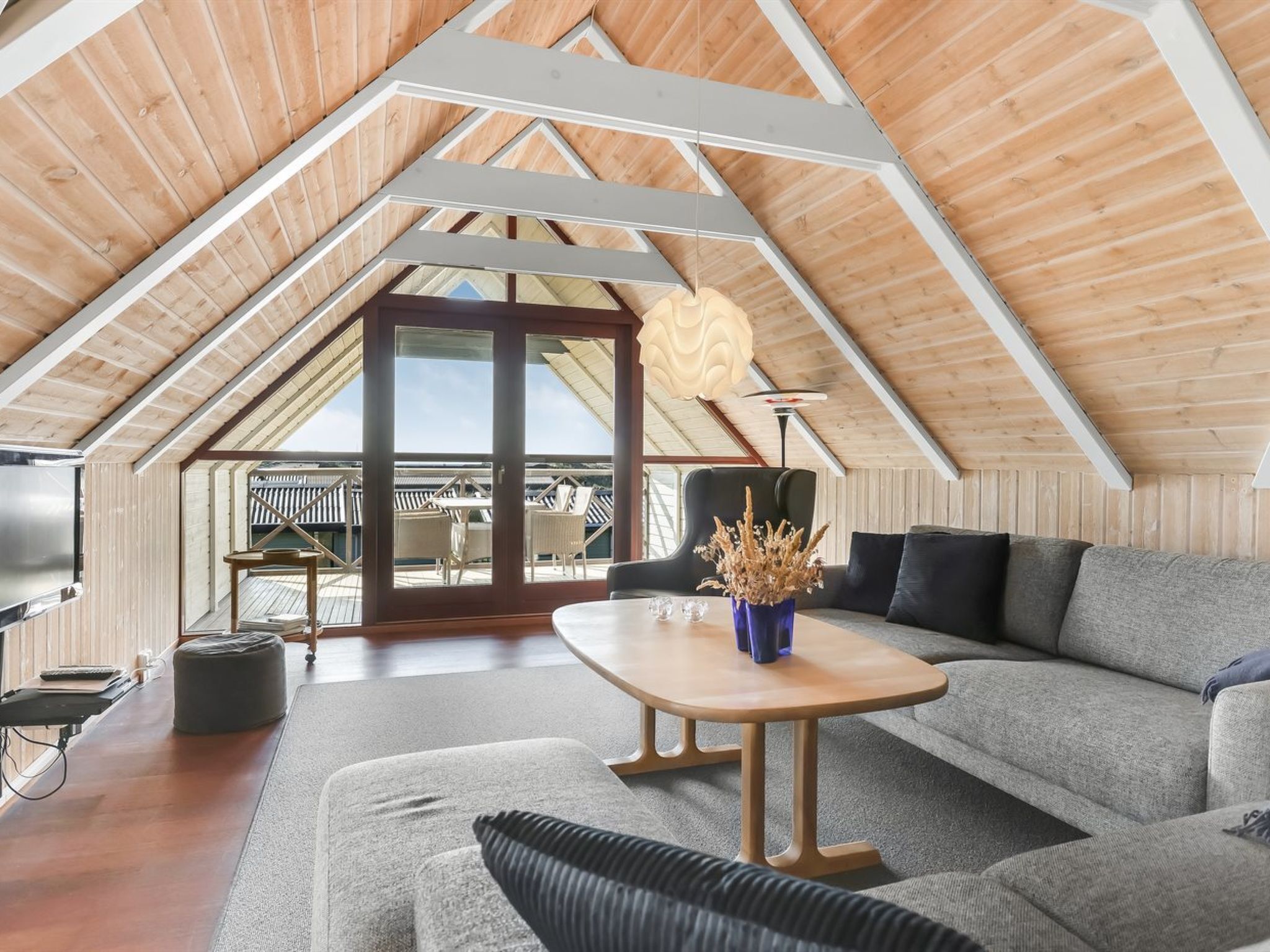 A modern loft with a sloped ceiling, a large sofa, a wooden table, and a view of the outdoors through a glass door.