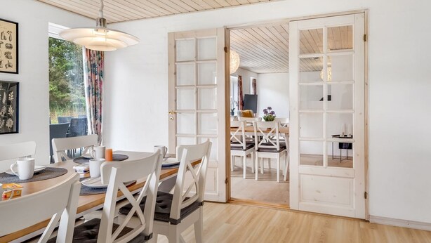 Un comedor moderno con techo de madera, paredes blancas y vistas a otra habitación por una puerta abierta.