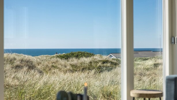 A view from a room with a large window overlooking a coastal landscape with dunes and the sea.