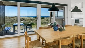 A modern interior with a wooden dining table, chairs, and a large sliding glass door leading to an outdoor area with a view of the beach and ocean.