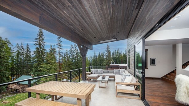 Ein geräumiger Balkon mit einem Holztisch und Bänken, einer Sitzecke mit Kissen und Blick auf eine bewaldete Landschaft.