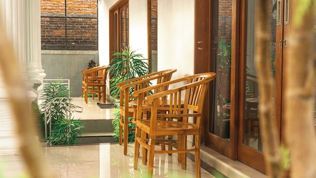 A hallway with wooden chairs and potted plants, leading to a door with a glass panel. - srinadi guest house (Buleleng)