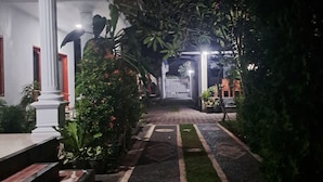 A pathway leading to a building entrance with a white column, steps, and a garden with plants and a bird. - srinadi guest house (Buleleng)
