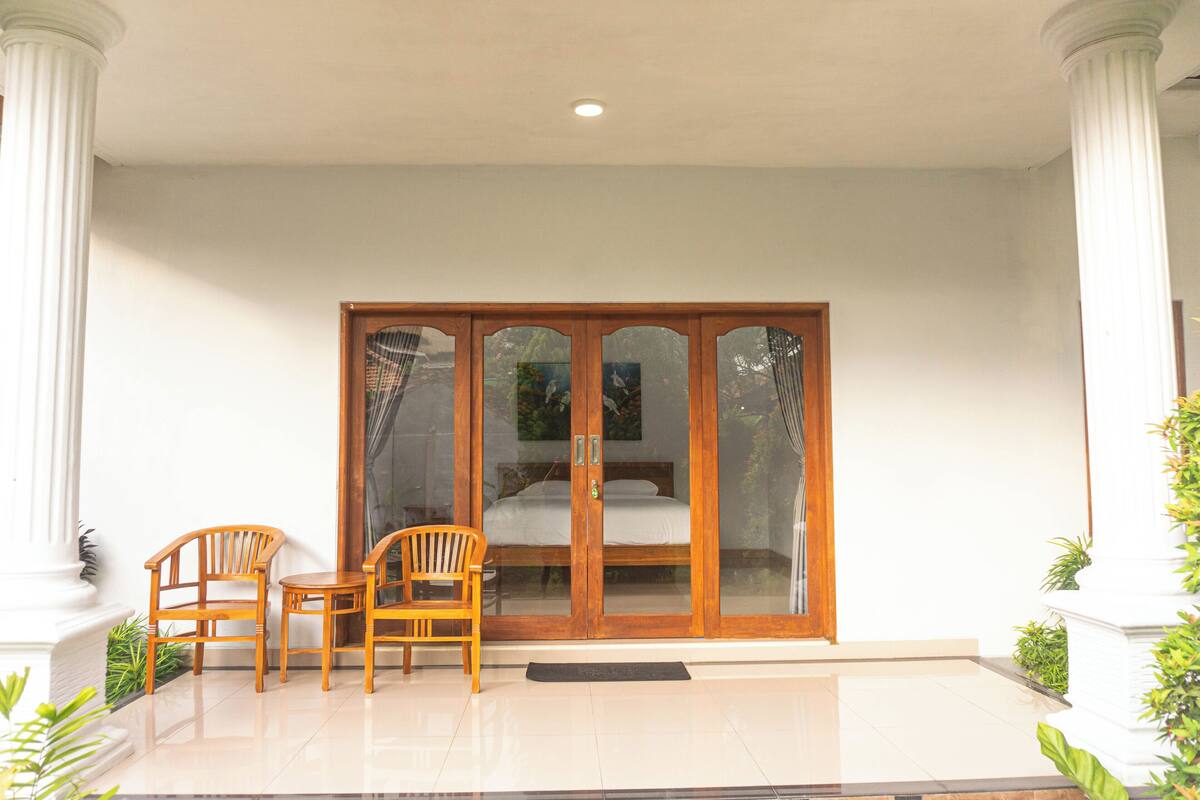 A room with a large sliding glass door leading to a bed, two wooden chairs, and a small table.