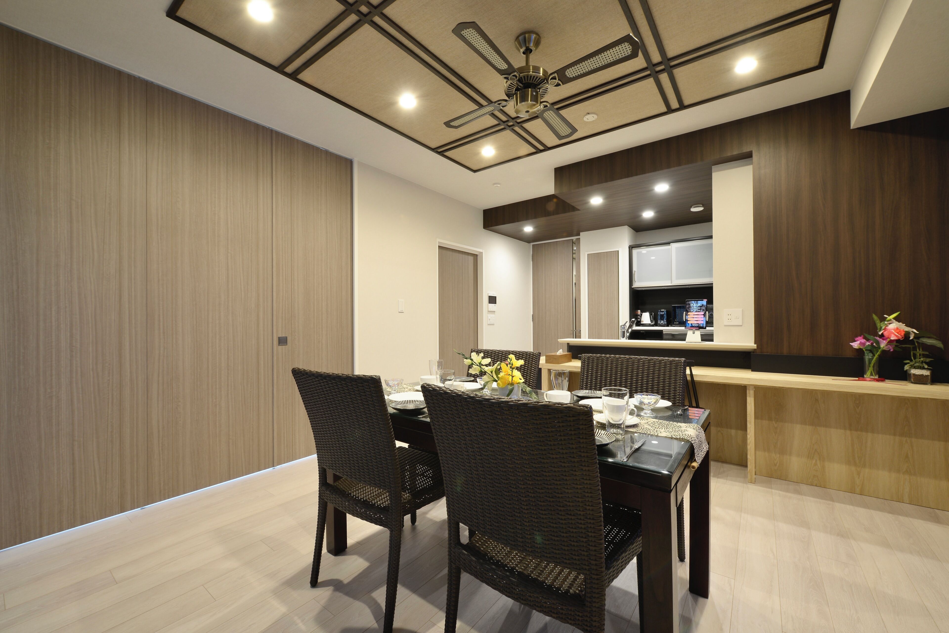 A modern dining area with a wooden table, dark wicker chairs, and a kitchen in the background.