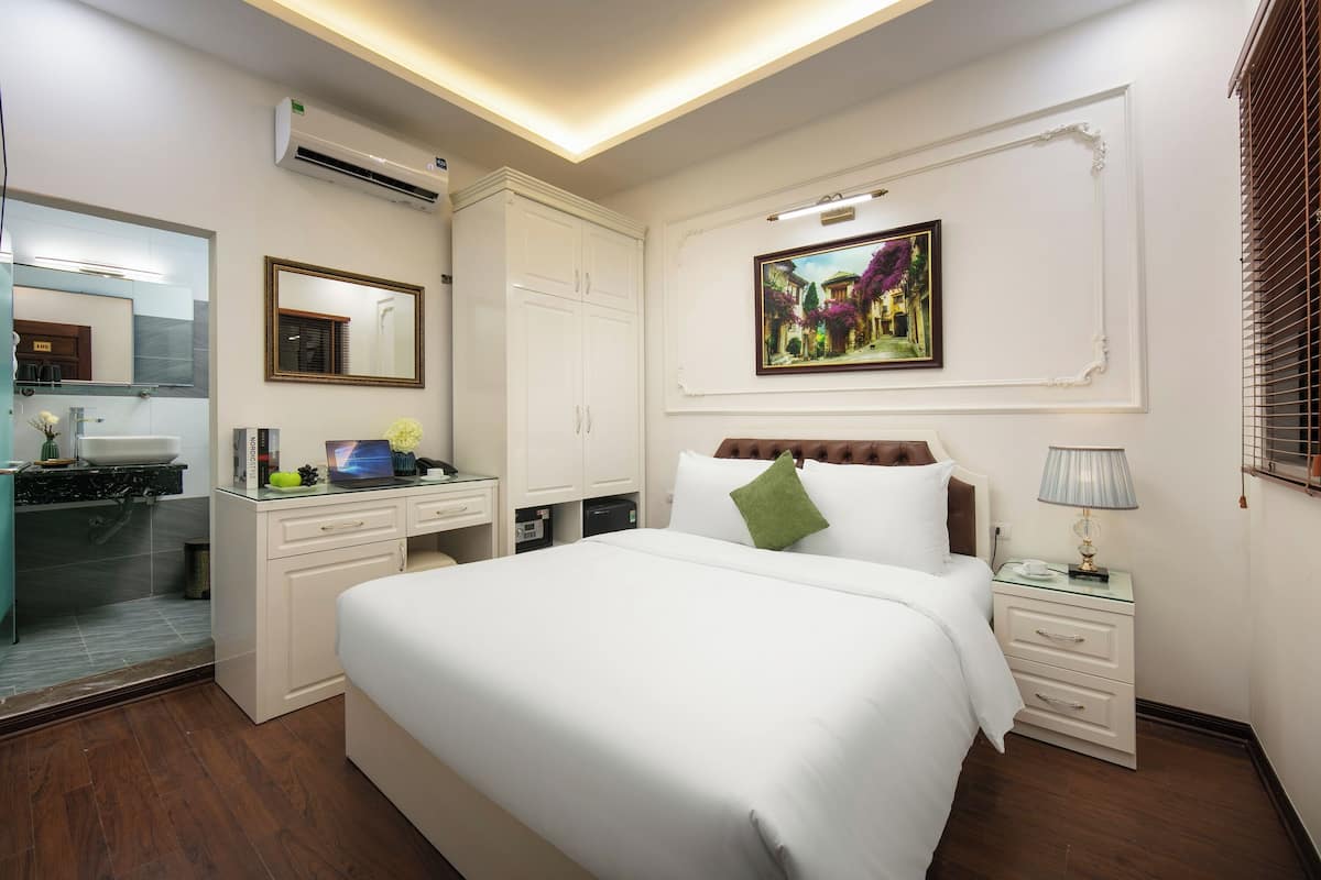 A hotel room with a large bed, a desk with a computer, a mirror, a white wardrobe, and a painting on the wall.