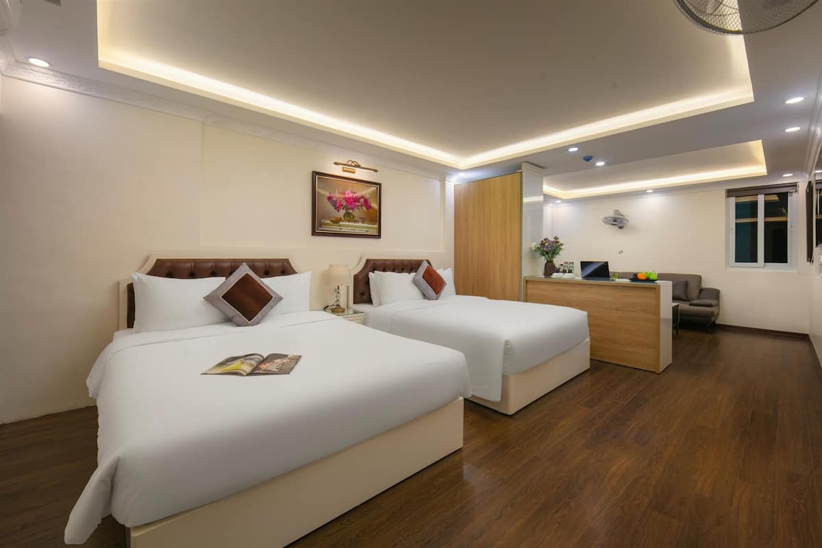 A hotel room with two beds, a desk, a chair, a fan, and a window.