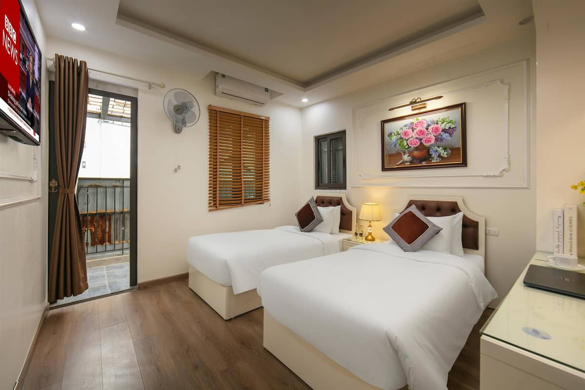 A hotel room with two beds, a TV, a fan, a painting, and a desk with a lamp.