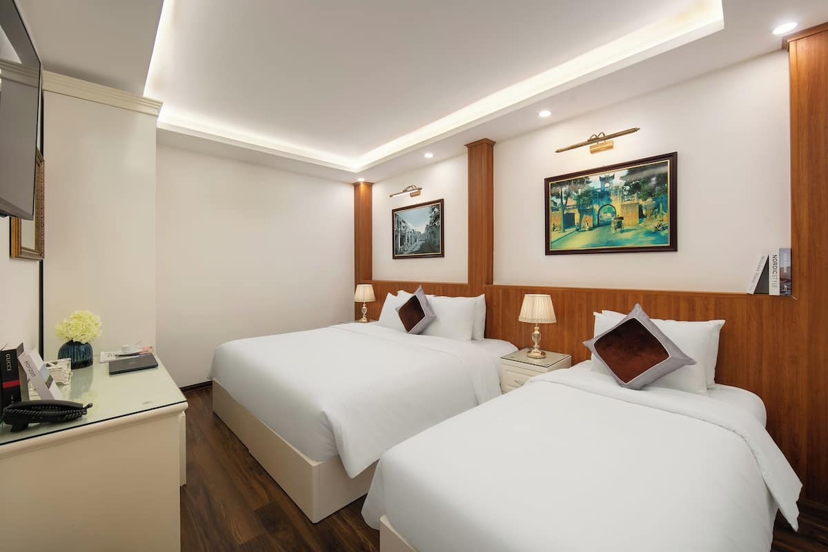A hotel room with two beds, a desk, and a nightstand. There are two lamps on the nightstand, a framed picture on the wall, and a small table with a vase of flowers.