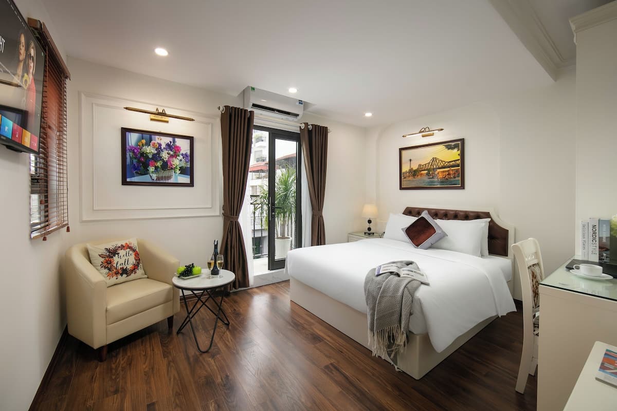 A hotel room with a bed, a chair, a small table with a lamp, a TV, and a window with curtains.