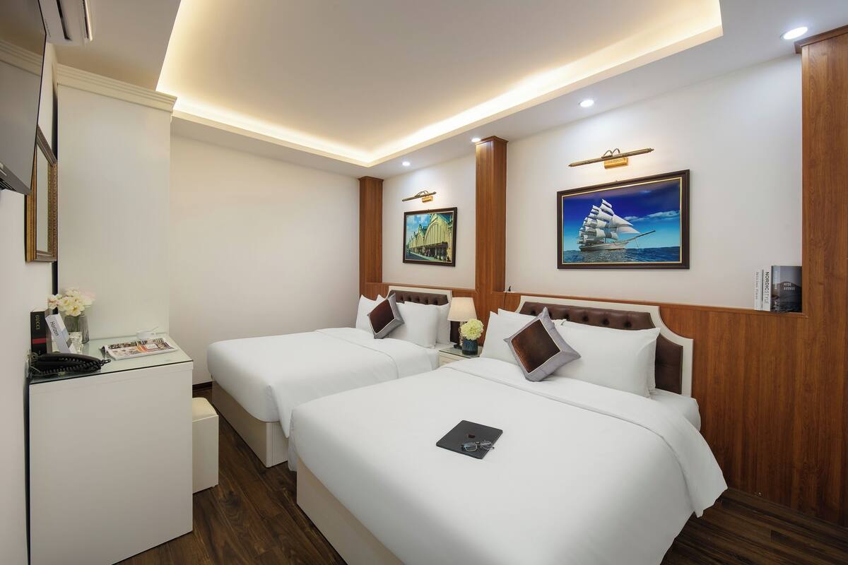 A hotel room with two beds, a desk, and a small fridge. There are framed pictures on the walls and a ceiling light.