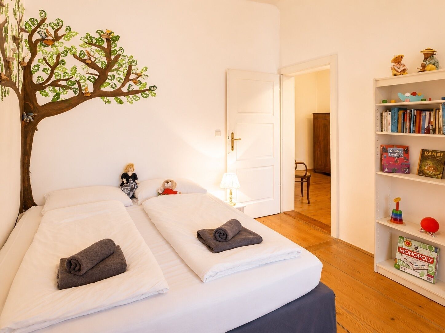 Ein Hotelzimmer mit zwei Betten, einem Bücherregal mit Kinderbüchern und einer Baum-Wandmalerei.
