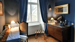 Ein Zimmer mit einem Bett, einem Holzstuhl, einem kleinen Tisch, einer Kommode und einem Fenster mit blauen Vorhängen.