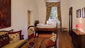 A cozy bedroom with a bed, a sitting area with a sofa and chairs, a wooden floor, and a large painting on the wall.