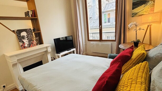 Een slaapkamer met een bed, een bureau, een stoel, een televisie, een raam met gordijnen en een schilderij aan de muur.