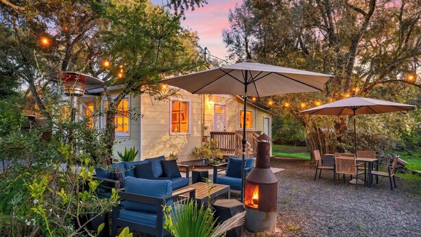 A cozy outdoor seating area with a fire pit, umbrellas, and a small building with windows. - Hobby Vineyard + Firepit + Outdoor Living + EV Charger | Grapevine Cottage (Glen Ellen)