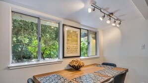 A dining area with a wooden table, chairs, and a bowl of snacks. There are two windows with a view of trees and a framed picture on the wall. - Hobby Vineyard + Firepit + Outdoor Living + EV Charger | Grapevine Cottage (Glen Ellen)