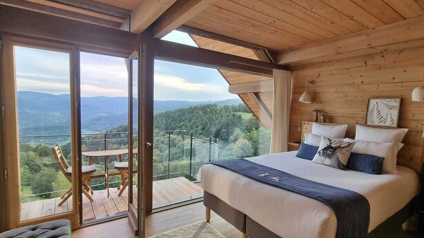 Une chambre à coucher avec un grand lit, une tête de lit en bois et une vue sur un paysage montagneux, visible à travers une porte-fenêtre coulissante.