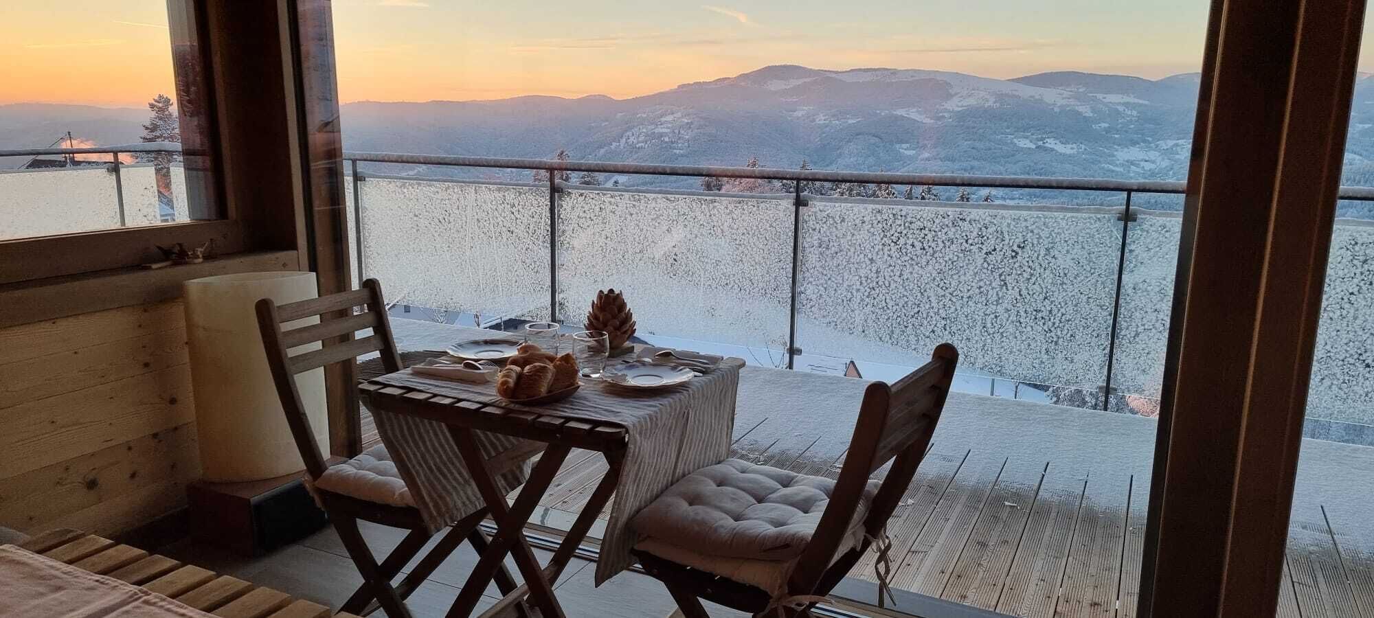A dining area with a table set for two, overlooking a snowy landscape with mountains in the distance.