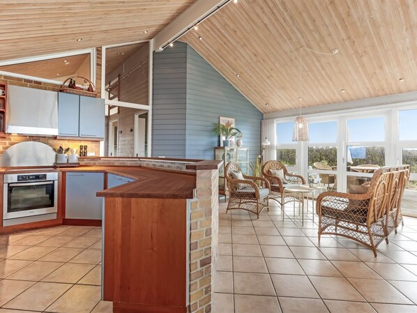 A spacious kitchen with a wooden island, a dining area with wicker chairs, and large windows offering a view of the outdoors. - "Aster" - 150m from the sea by Interhome (Hirtshals)