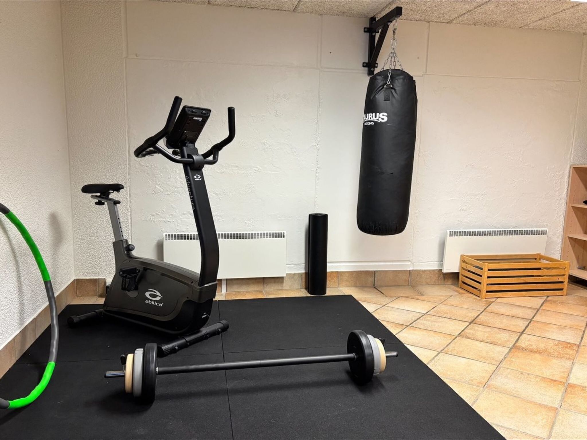 Ein Heimfitnessraum mit einem Crosstrainer, einem Sandsack und einer Langhantel auf einer schwarzen Matte.