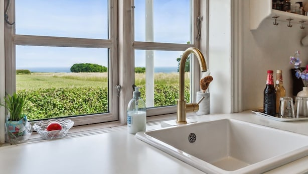 Eine Küche mit einem großen Fenster, einem weißen Spülbecken und Blick auf eine grüne Landschaft.
