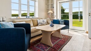 Ein geräumiges Wohnzimmer mit einer Couch, Sesseln, einem Couchtisch und einem Bücherregal. Große Fenster sorgen für natürliches Licht und einen Ausblick ins Freie.