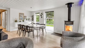 Uma sala de estar moderna com lareira, uma área de jantar com mesa e cadeiras brancas, e vista para o exterior através de grandes janelas.