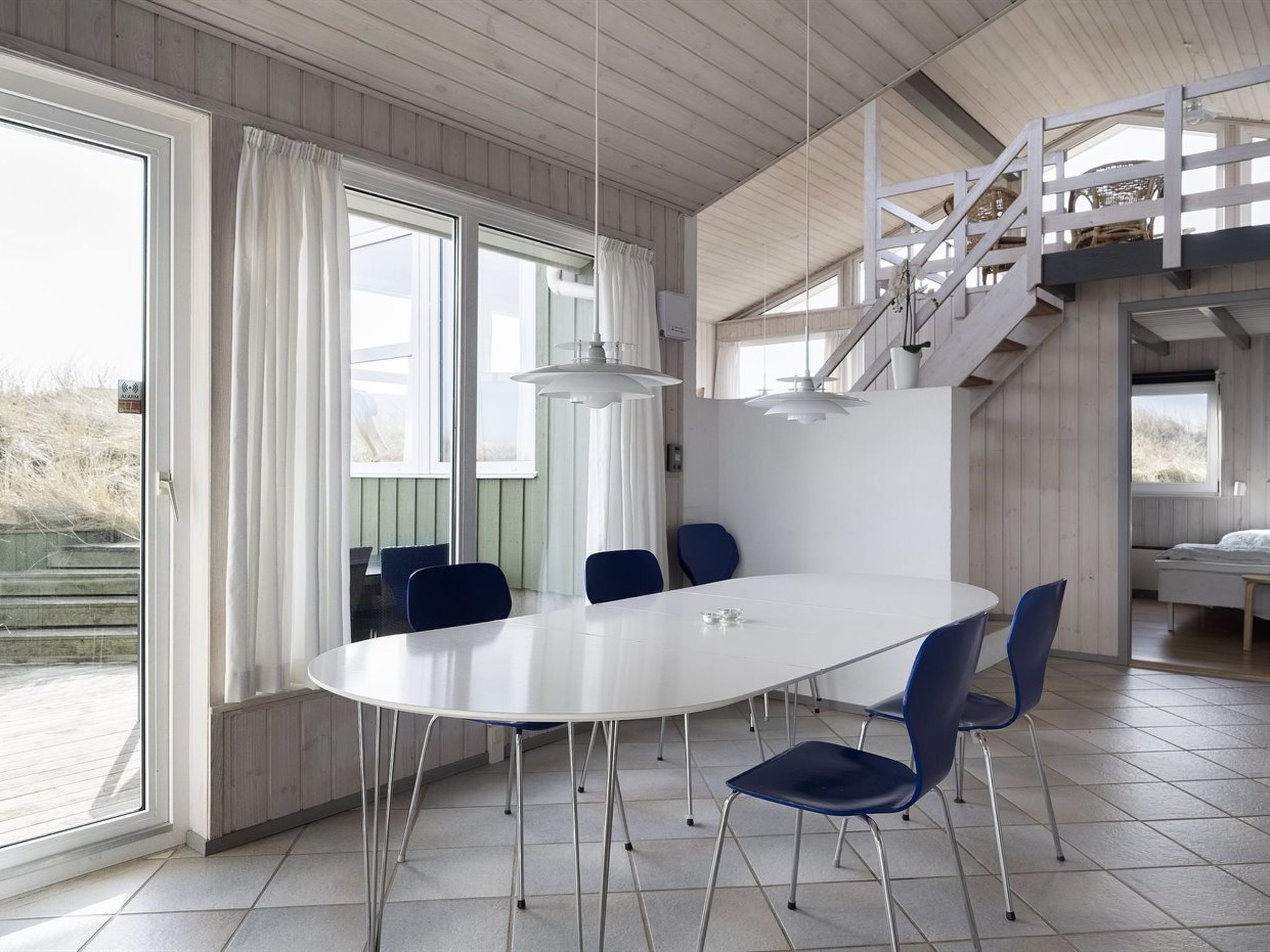 Ein moderner Essbereich mit einem weißen Tisch und blauen Stühlen, großen Fenstern mit Vorhängen und einer Treppe zu einer oberen Ebene.