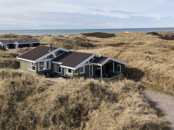 Una fila di case sulla spiaggia, con un sentiero sabbioso che le conduce, circondate da dune e il mare sullo sfondo.