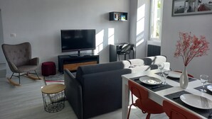 Een moderne woonkamer met een flatscreen televisie, een bank, een eettafel gedekt voor twee personen en een rode fauteuil.
