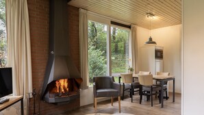 Un salon moderne doté d’une cheminée, d’un plancher en bois, d’un coin repas avec des chaises et une table, et de grandes fenêtres offrant une vue sur la verdure.