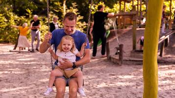 A man and a child on a swing in a park with sand on the ground and wooden structures in the background.