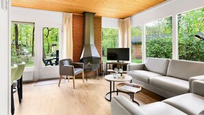 Una sala de estar moderna con chimenea, sofá y televisor, ventanales amplios con vistas a zonas verdes.