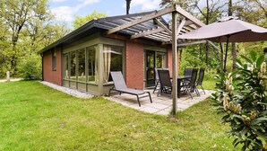 En glasindkapslet terrasse med et bord og stole, omgivet af en murstensbygning og en træpergola.