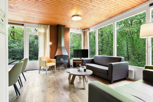 A modern living room with a fireplace, comfortable seating, and large windows offering a view of the forest.