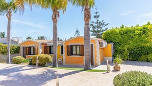 A two-story house with a tiled roof, a small porch, and a garden with palm trees and shrubs. - 4 Bedroom Villa in Pozzallo (Pozzallo)