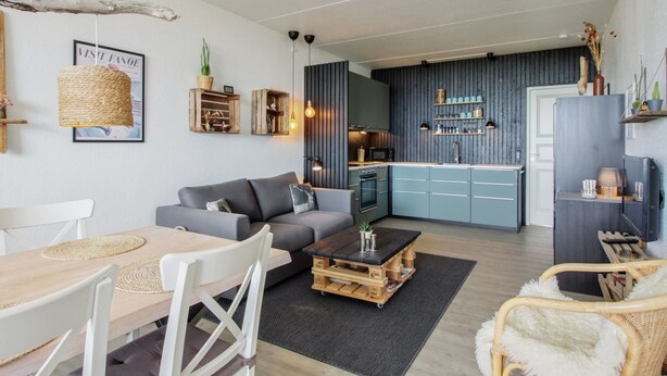 Uma sala de estar moderna com um sofá cinzento, mesa de centro de madeira e cadeiras de vime. Uma cozinha com armários azuis e uma área de refeições com mesa e cadeiras de madeira são visíveis ao fundo.