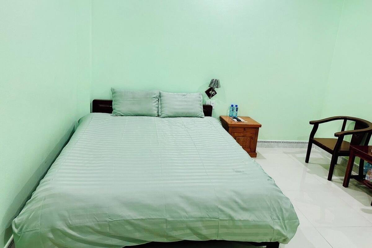Un lit simple avec une couvre-lit verte et deux oreillers, une table de chevet en bois avec une lampe et une bouteille, et une chaise en bois.