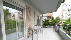 Uma varanda com mesa e cadeiras de vidro, plantas em vasos e vista dos edifícios ao redor.