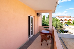 A balcony with a table and chairs, a sliding glass door, and a view of a building with arches and trees. - Bonvie Apartments in Center 12 B6 (Novalja)