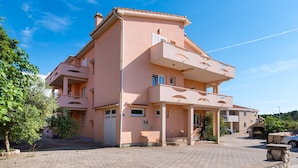 A multi-story building with a flat roof, a balcony on the second floor, and a driveway leading to a gated entrance. - Bonvie Apartments in Center 12 B6 (Novalja)