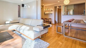 A modern living room with a sofa, a dining table, and a kitchen area with a refrigerator and shelves.