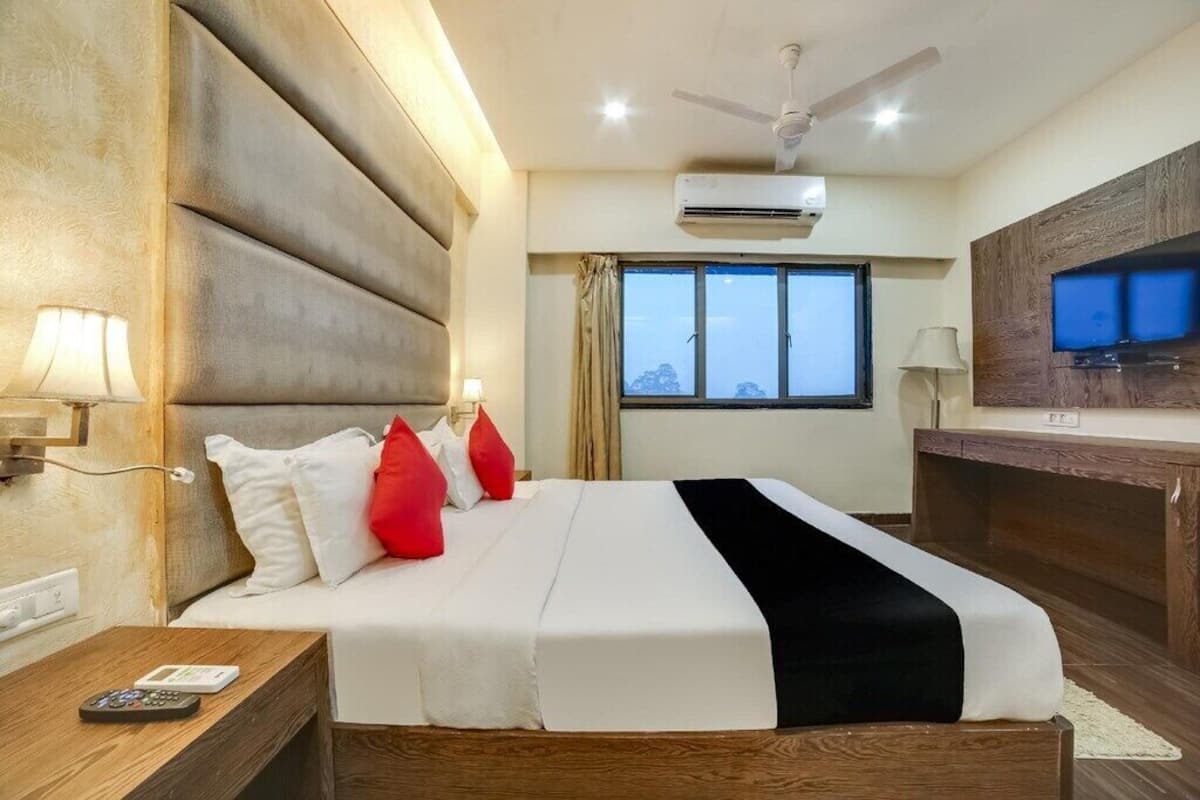 A hotel room with a large bed, a flat-screen TV, an air conditioning unit, and a city view through the window.