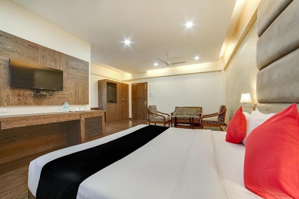 A hotel room with a bed, a TV mounted on a wooden panel wall, a desk with a chair, and a seating area with two chairs.