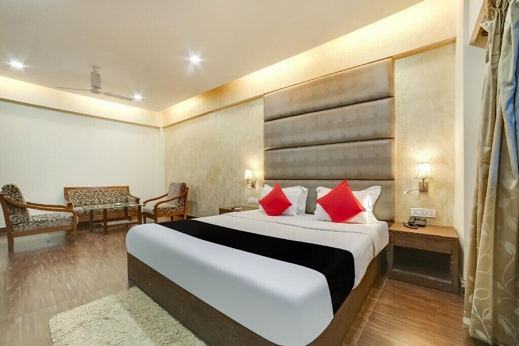 A modern hotel room with a large bed, a sofa, two chairs, a nightstand with a lamp, and a ceiling fan.