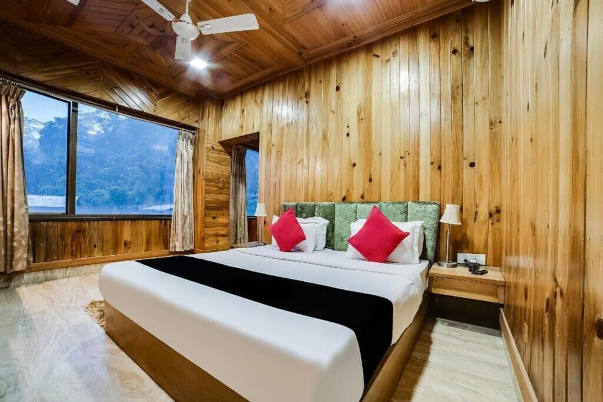 A wooden-paneled room with a large bed, a nightstand, a lamp, and a window with a view of trees.