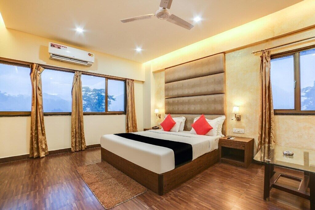 A hotel room with a large bed, a ceiling fan, an air conditioning unit, a nightstand, a desk, and large windows with curtains.